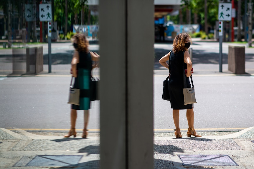 A woman wearing a mask standing on the side of a street, on a sunny day.