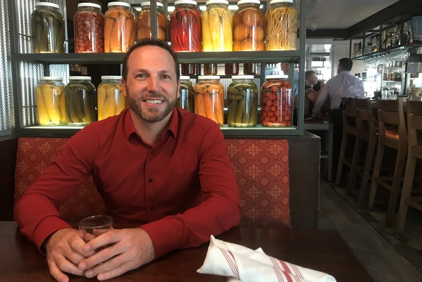 A man in a red button down shirt sits in a booth at a restaurant with jars of pickles on shelves behind him.