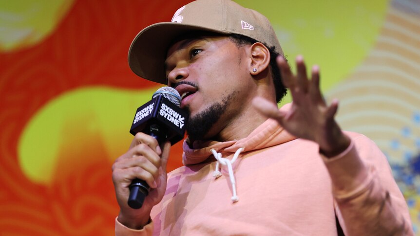 Chance The Rapper speaks into a microphone and holds up a hand. He wears a brown baseball cap and pink sweater.