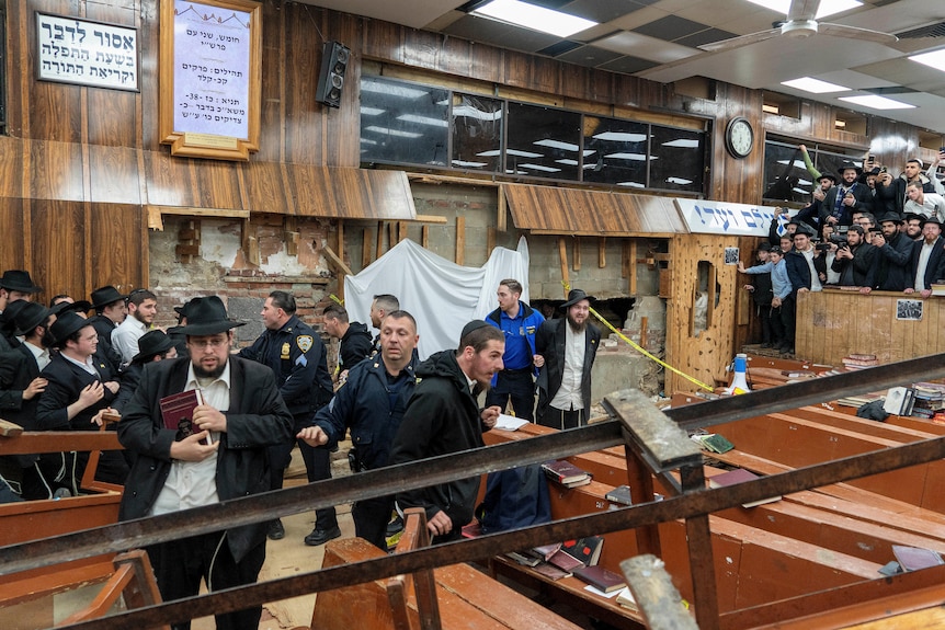 A wide shot inside a synagogue. Police tussle with Jewish worshippers near a broken wall, as other worshippers watch on nearby