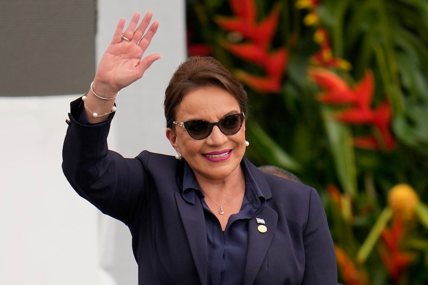 A middle-aged central American woman in a navy outfit and sunglasses smiles and waves to someone in the distance.