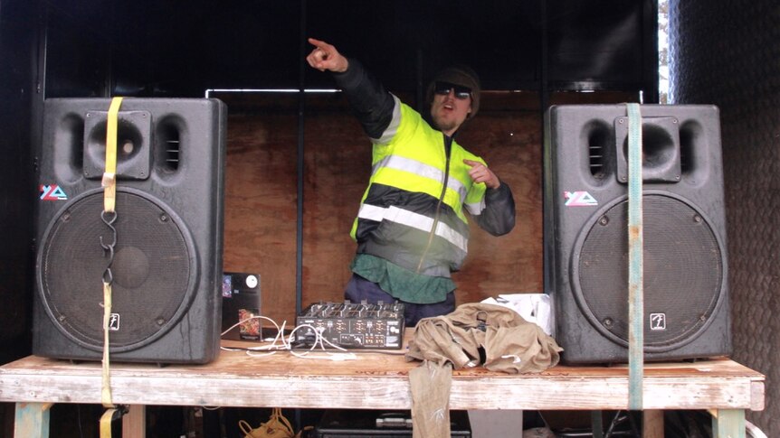 A DJ pumps music out of a mobile booth with speakers as volunteers plant trees on a farm