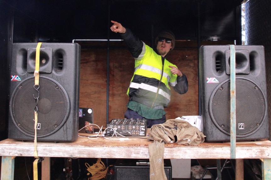A DJ pumps music out of a mobile booth with speakers as volunteers plant trees on a farm