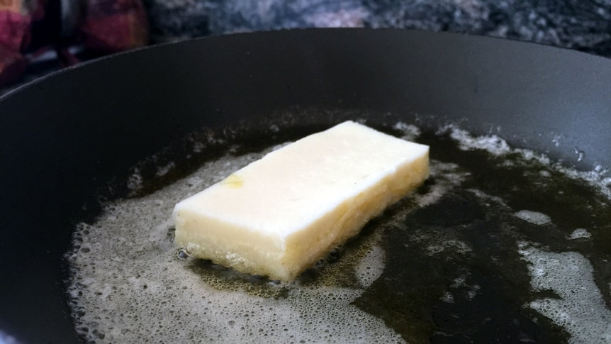 a small off-white rectangle sits in a pool of butter in a black frying pan