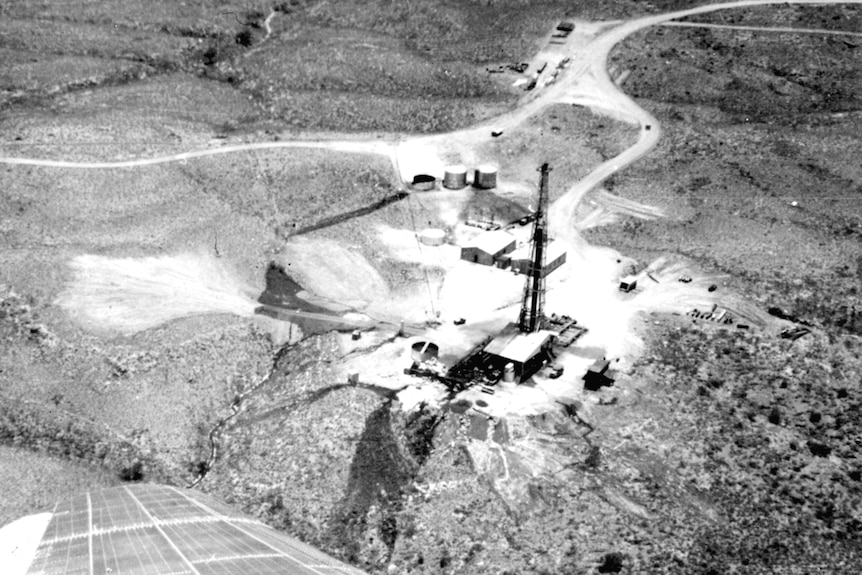 A black and white aerial photograph looking down at a lone oil rig on a rocky range.