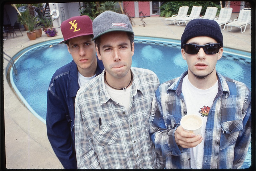 Two men wearing baseball caps stand close to man in beanie and sunglasses, all three pose in front of small round outdoor pool.