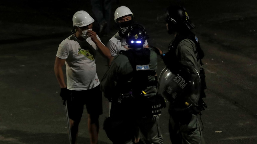 Men in white t-shirts and carrying poles talk to riot police in dark green jumpsuits in Yuen Long, Hong Kong