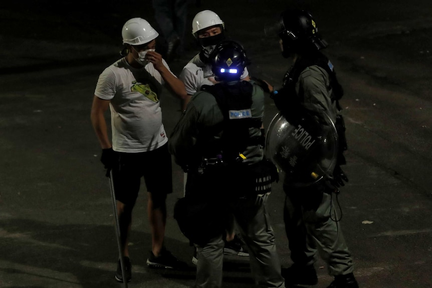 Men in white t-shirts and carrying poles talk to riot police in dark green jumpsuits in Yuen Long, Hong Kong