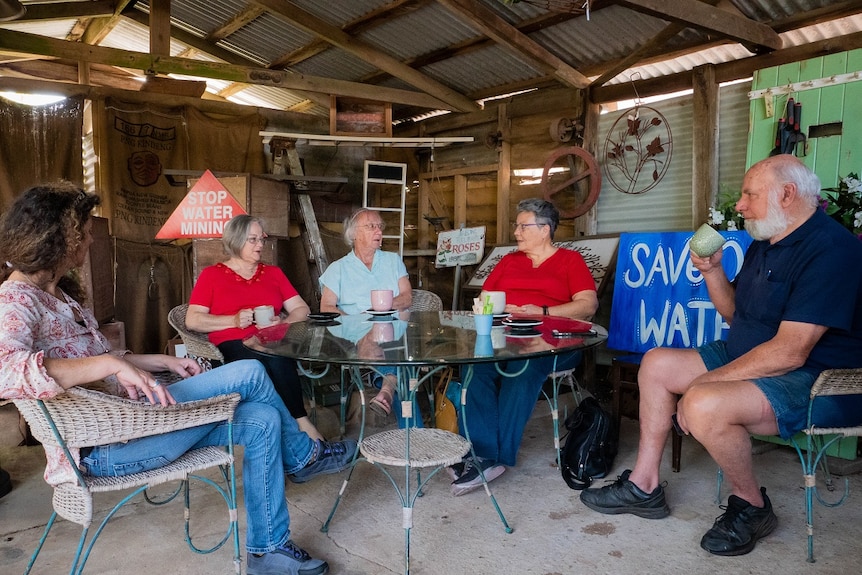 three women and a man sit around a table in a tin shed having cups of tea. anti-water mining signs are in the background