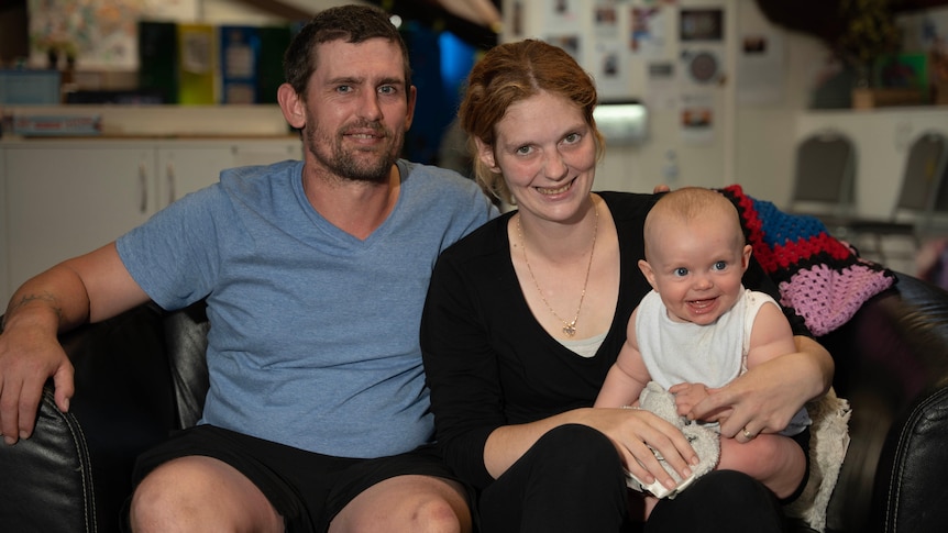 A man, woman and baby sit on a couch and smile at the camera.