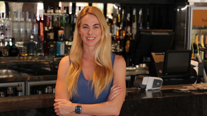 Bree stands behind the bar with her arms folded, smiling.