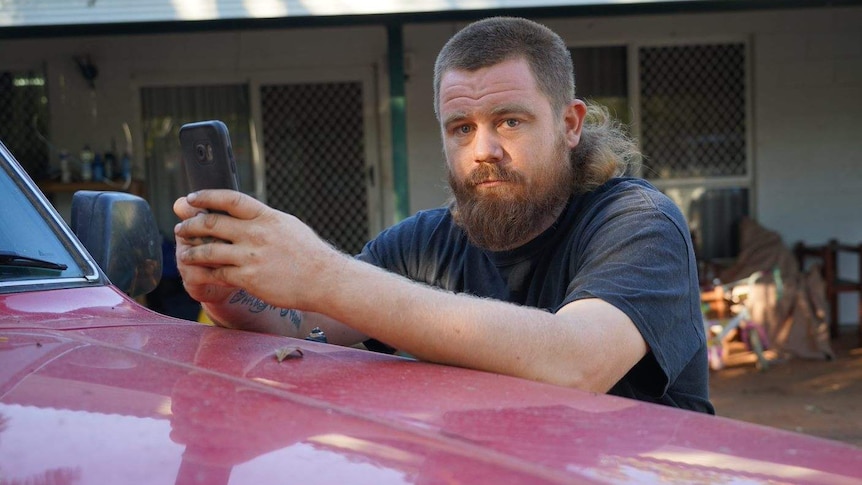 A man with a mullet leaning on the bonnet of a car holding a phone and looking unhappy