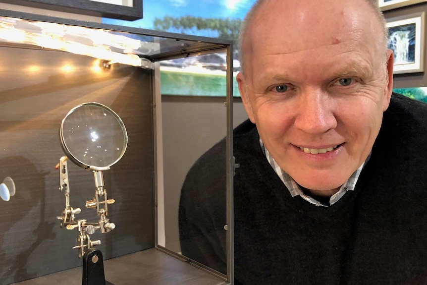 Artist Wayne Malkin next to a glass box with magnifying glass, and matchstick with a tiny painting on it