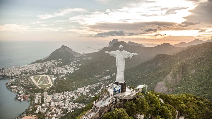large christ the redeemer statue overlooking forests, mountains and coastal city, with setting sun in the background