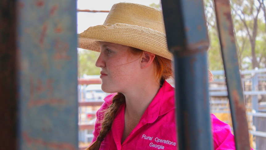 A teenage girl with a pink shirt and wide brim hat stands beside a cattle crush.
