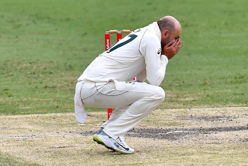 Nathan Lyon holds his head in his hands while squatting down next to the wickets