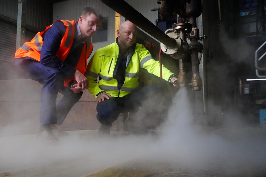 Two men in high vis squad next to a tap and pipe watching steam spurt out the end in a factory