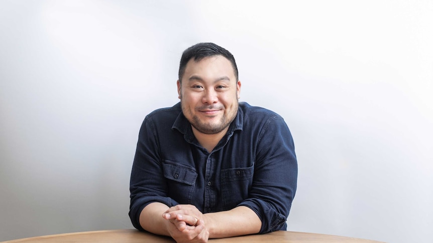 The chef David Chang wearing a blue shirt sitting at a bare table