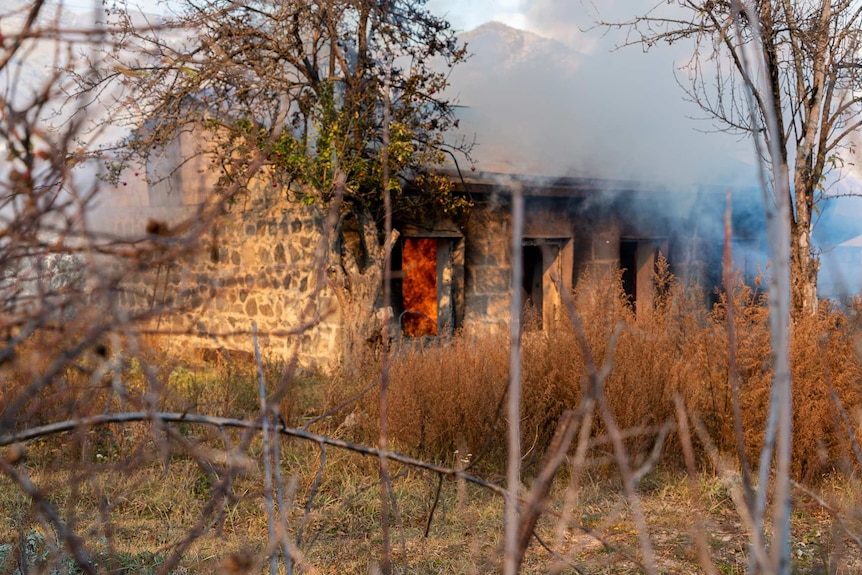 A stone house in a field with orange flames coming through the windows and doors