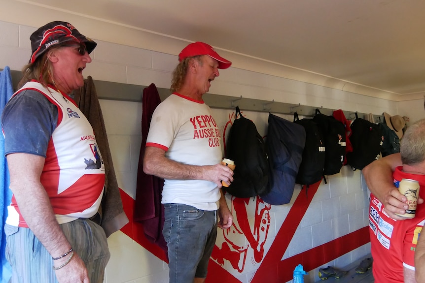 Two men in Yeppoon Swans shirts smiling and singing in a white locker room.