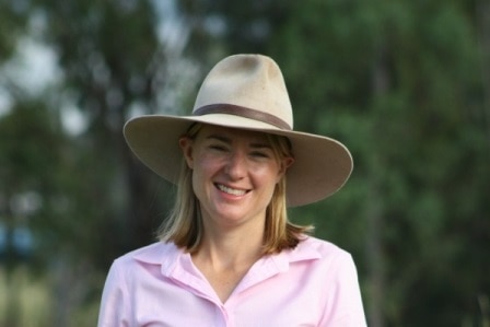 Peta Ward standing in a pink work shirt with an akubra with green trees in the background.