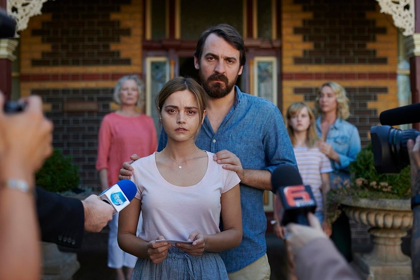 A woman and man stand in front of their house in the spotlight of cameras and microphones