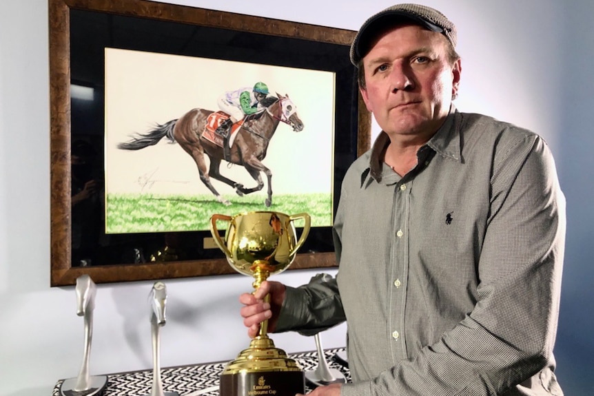 A man holds a gold cup, with a framed picture of a horse and jockey on the wall behind him.