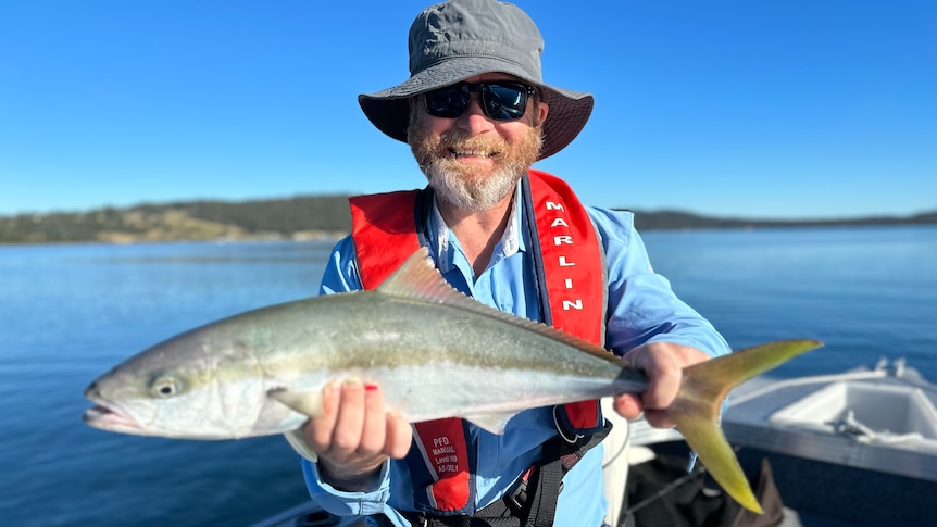 ABC Statewide Mornings presenter Leon Compton sitting in a boat on the water holding a kingfish 
