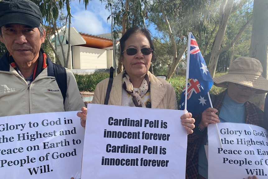 Several people stand in line holding signs supporting George Pell.