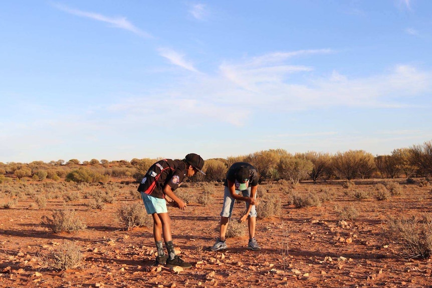 Two young boys bend over red, rocky ground.