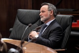 A man with grey hair and a grey beard is wearing a suit. He's leaning back in his chair and appears to be watching something.