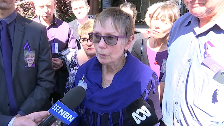 An older woman wearing a purple scarf and flanked by supporters wearing purple, speaks to media-branded microphones outside