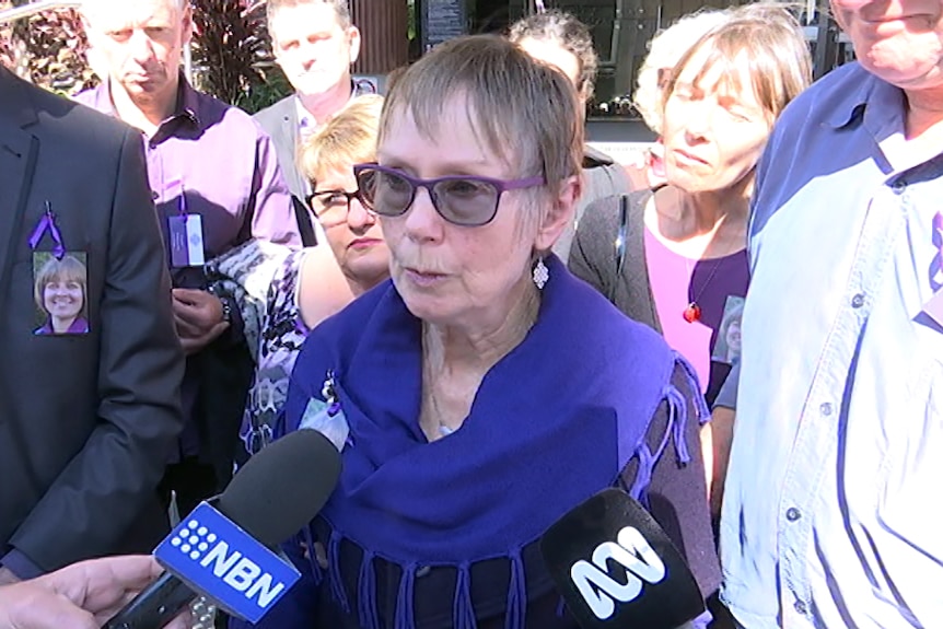 An older woman wearing a purple scarf and flanked by supporters wearing purple, speaks to media-branded microphones outside