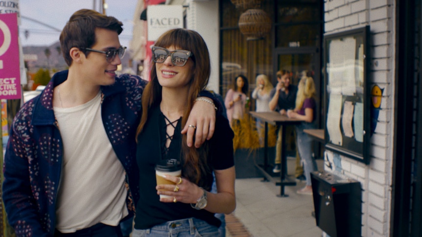 Anne Hathaway and Nicholas Galitzine in character outside, arms slung over shoulder, holding coffee cup, wearing sunglasses