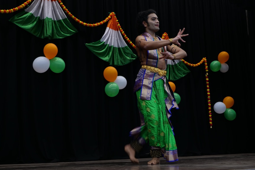 Indian man in vibrant green, gold and blue costume performing a dance on stage