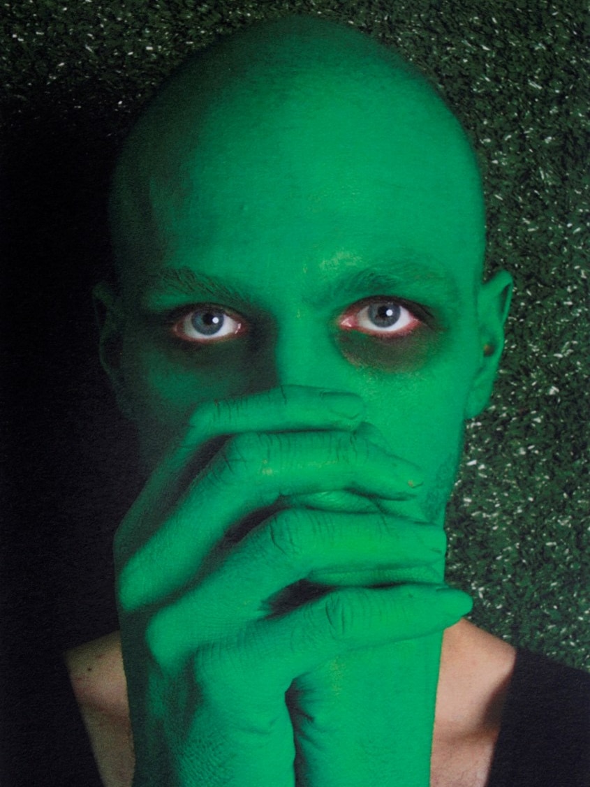 A bald man with his hands covering the bottom half of his face, his face and hands are painted in a bright green