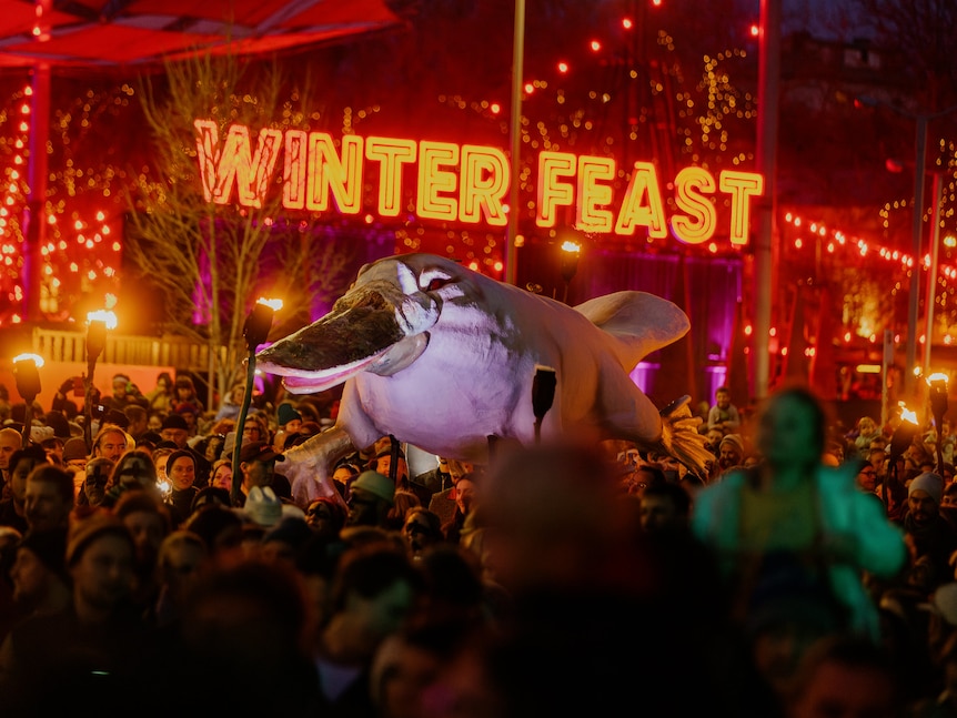 Crowd surrounds a platypus sculpture being carried past illuminated sign.