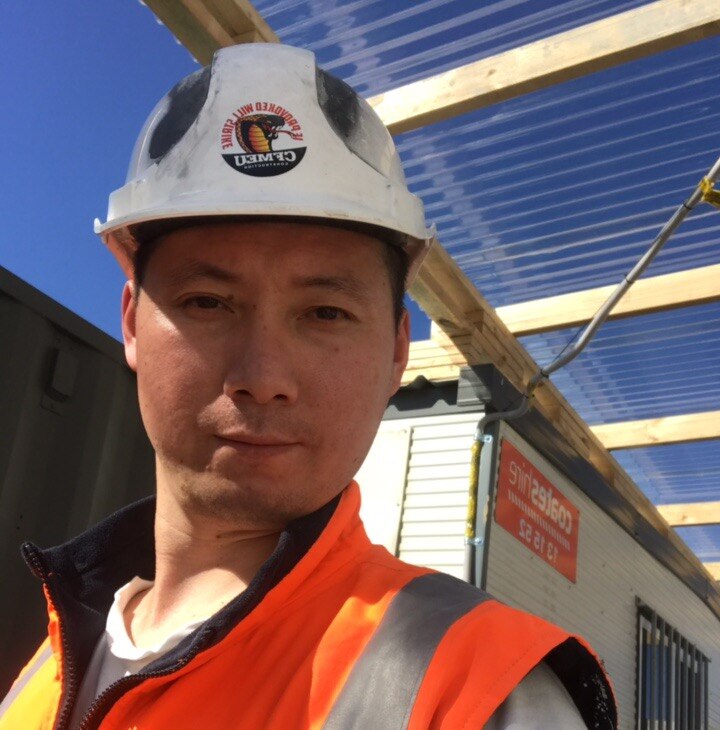 A man smiles into the camera. He is wearing a hard hat and is at a work site.