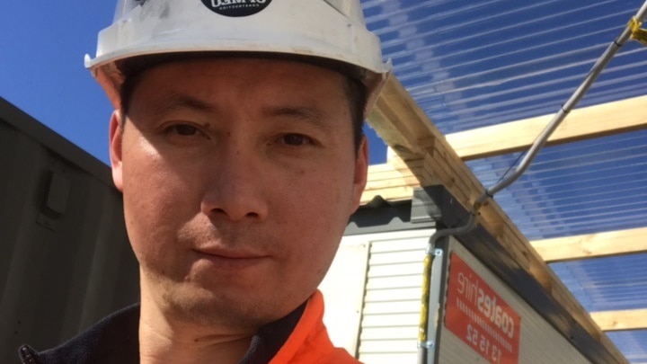 A man smiles into the camera. He is wearing a hard hat and is at a work site.