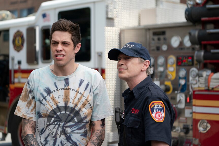 In front of a fire truck stands older uniformed man and young man in tie-dye t-shirt with unimpressed expression.