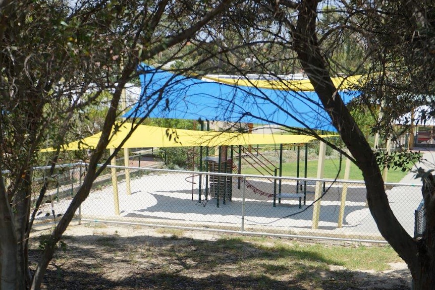 A wide shot of a school playground under blue and yellow shade sails with trees in the foreground.