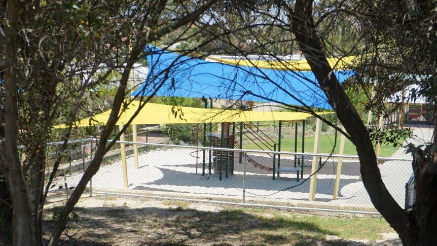 A wide shot of a school playground under blue and yellow shade sails with trees in the foreground.
