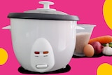A basic white rice cooker, next to a measuring cup of rice and some veggies, on a pink background.