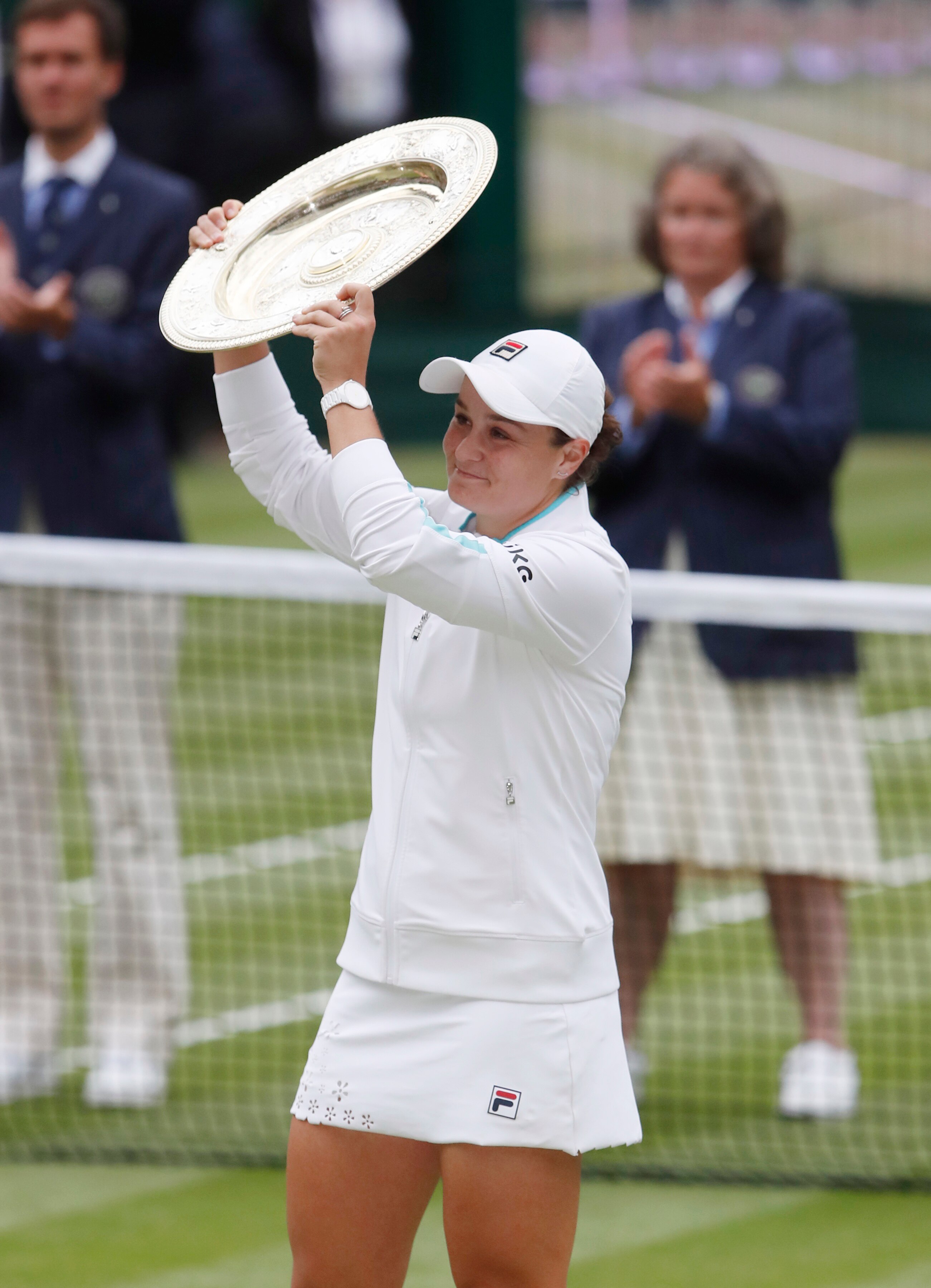 A young woman in white holds a plate trophy above her head on a grass court with people clapping behind.