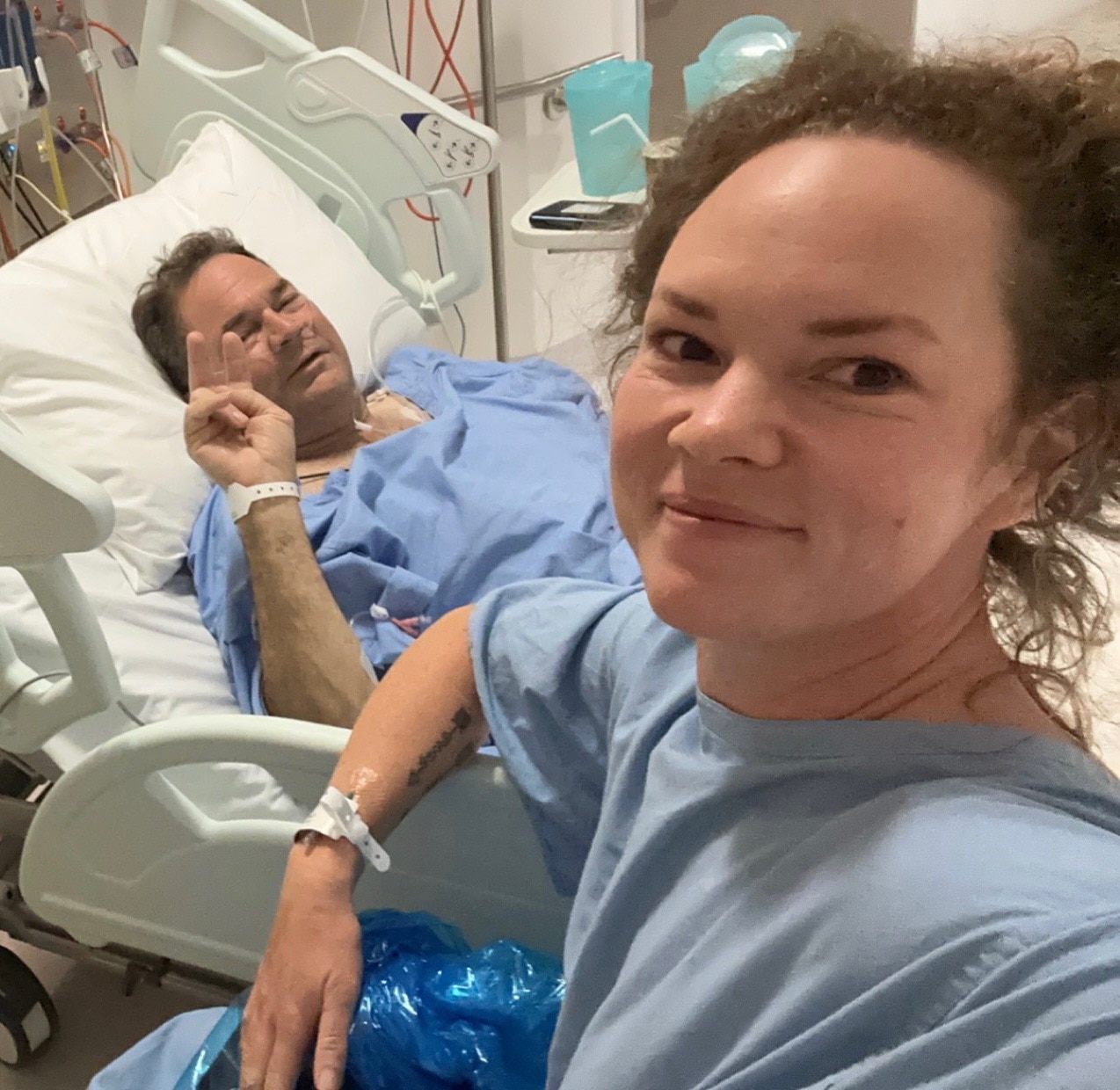 A man and woman in hospital scrubs. The man is lying down in bed holding two fingers in a peace sign.