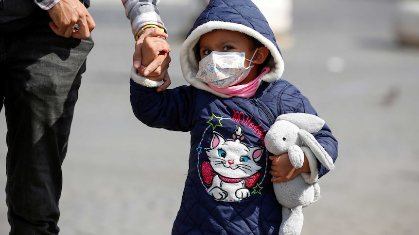A child holding a parent's hand and clutching a toy rabbit wears a face mask.