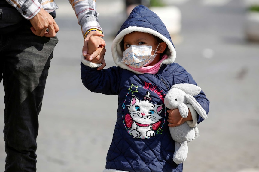 A child holding a parent's hand and clutching a toy rabbit wears a face mask.