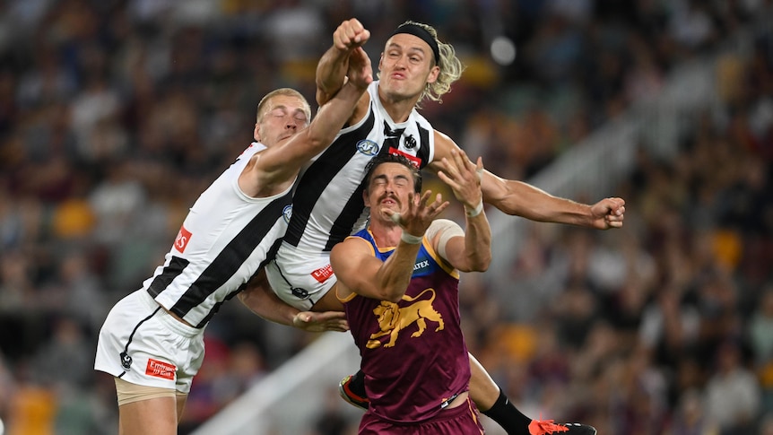 A Collingwood player leaps high and punches the ball clear as he is one of three players grimacing in a contest.