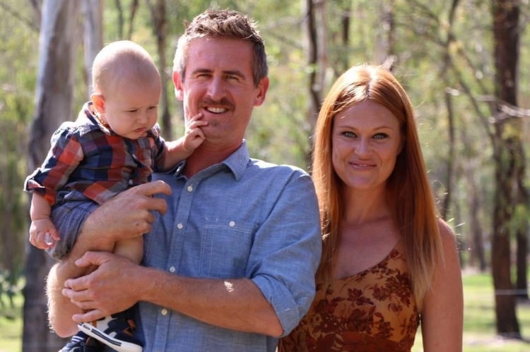 A woman and a man. The man is holding a baby on a sunny day.