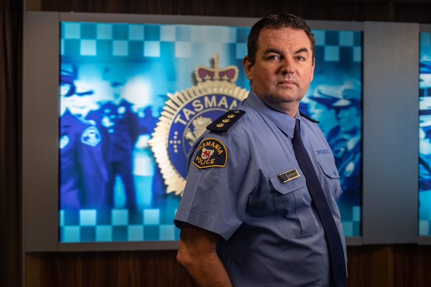A police officer in uniform stands in front of a back drop reading: Tasmania Police